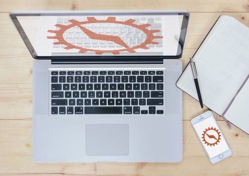 The Columbia Gadget Works gear logo on a laptop screen and a phone, on a wooden desk beside a notebook and pen